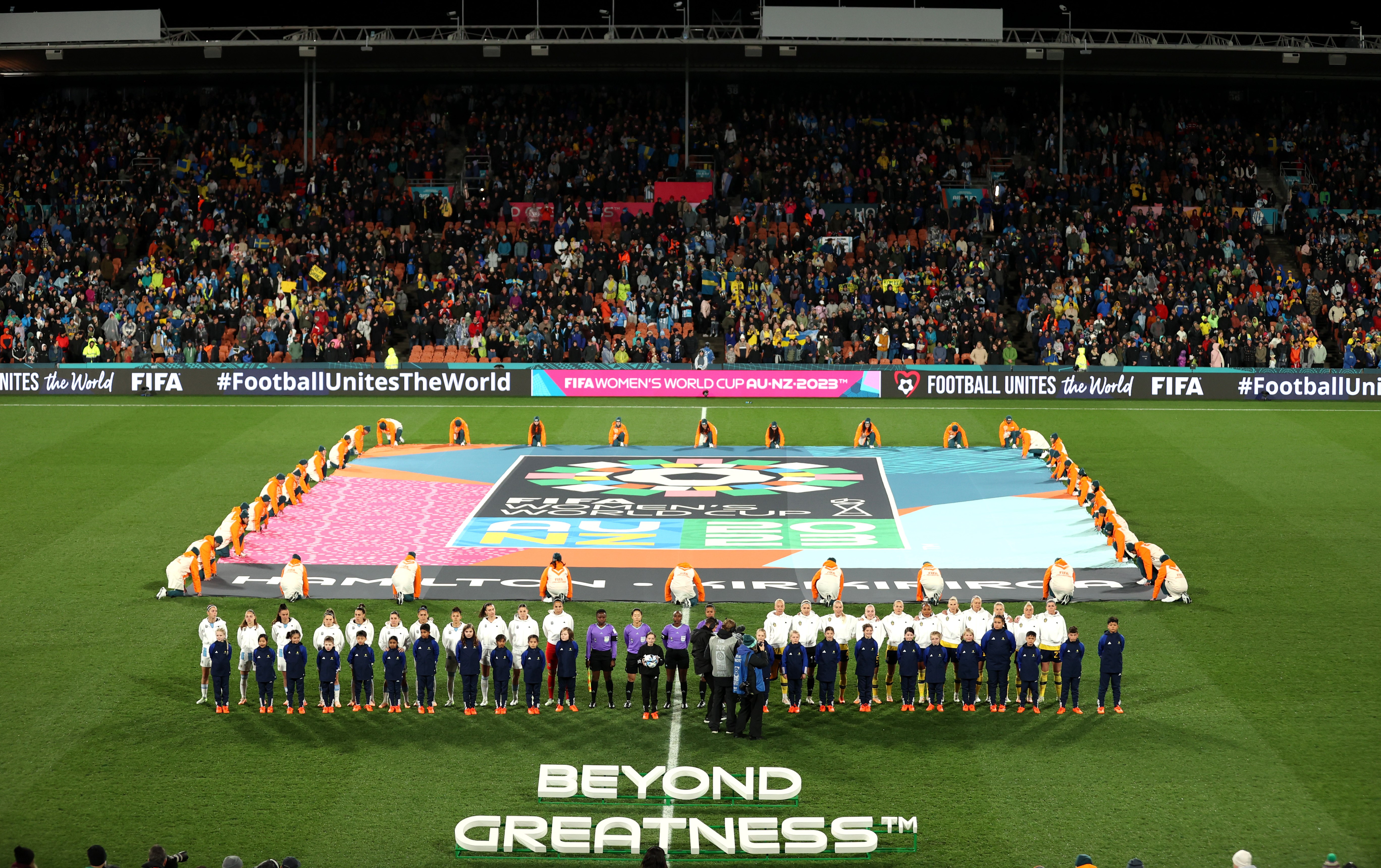 A group of people kneeling down around a big FIFA Women's World Cup flag laid out on the field with a group of people standing in front facing the crowd.