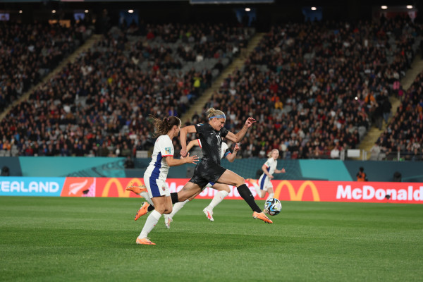 A woman footballer in action shot of kicking the football while the opposition is running behind in competition to get the football.