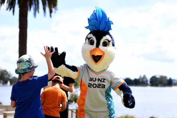Penguin mascot dressed in a football shirt that has AU-NZ 2023 written on it, giving a young kid a high-five