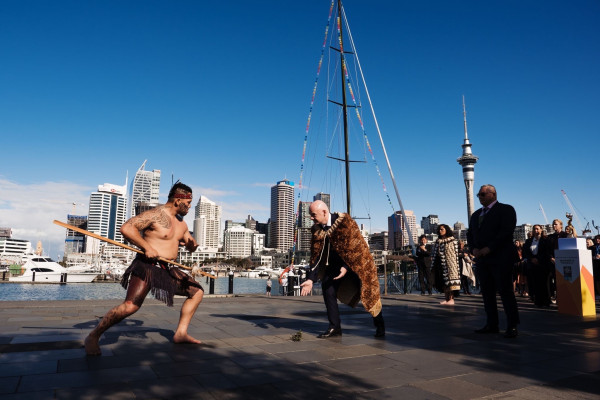 One man performing a traditional Māori stance, holding a Māori stick. Another man wearing a Māori cloak bending down to receive a gift from the ground and a group of people standing behind him. 