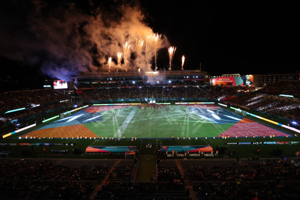 A night image of a football stadium with the field pitch painted in colours of green, orange, red, and blues and fireworks displaying on top of the grandstand.